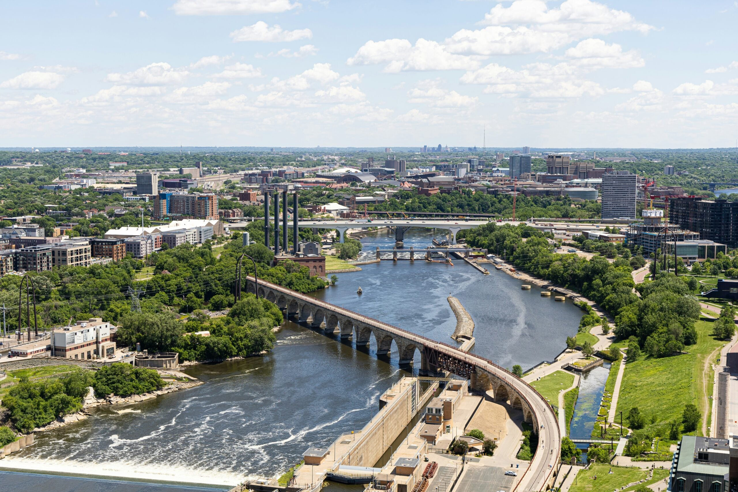 Aerial view of a river with a long stone arch bridge crossing it, surrounded by green trees and city buildings under a blue sky with scattered clouds.