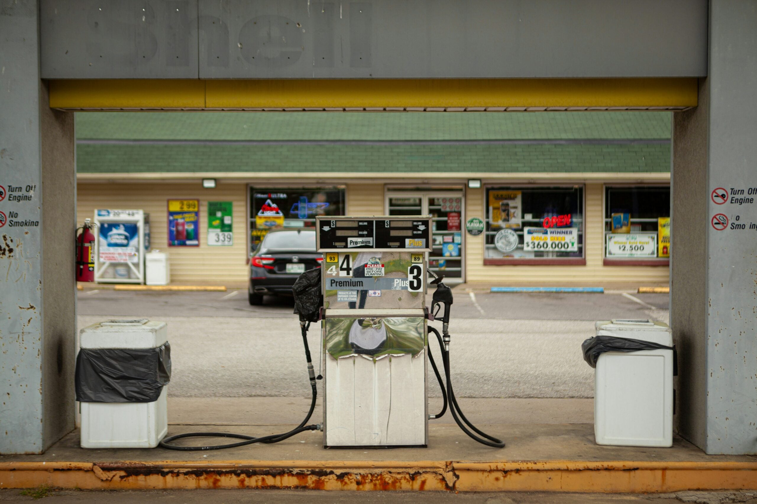 A single gas pump sits between two trash cans at an outdoor station, with a convenience store and parked cars visible in the background. The faded Shell sign is barely visible above.
