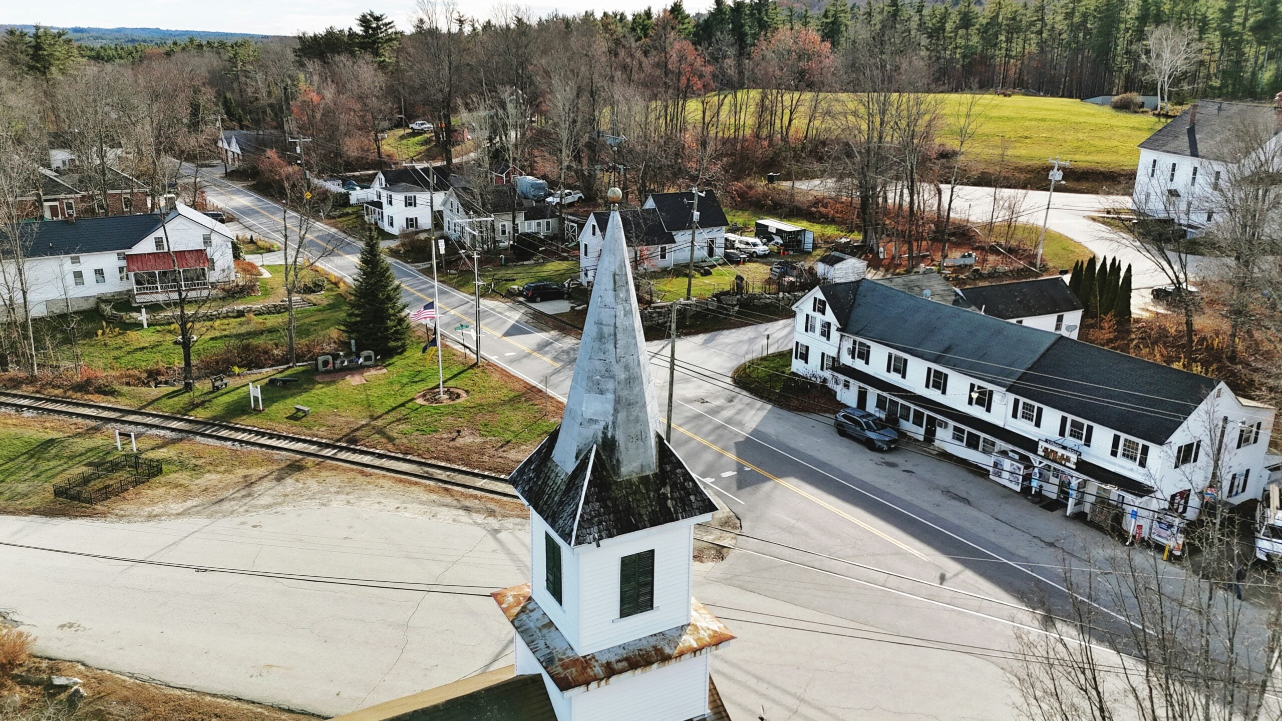 Aerial view of a small rural town with a church steeple in the foreground, intersecting roads, railroad tracks, white houses, and trees with sparse autumn foliage.