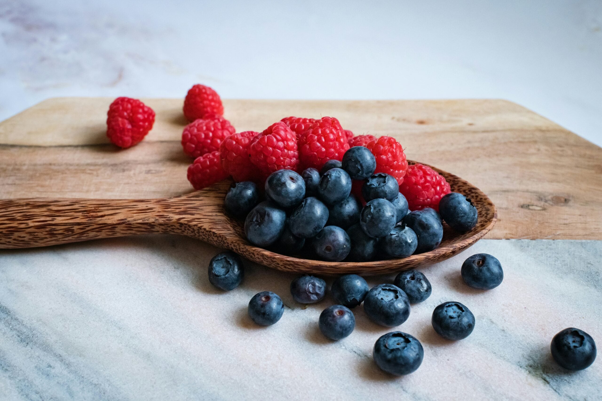 A wooden spoon filled with fresh blueberries and raspberries rests on a wooden cutting board, with some berries scattered on a marble surface.