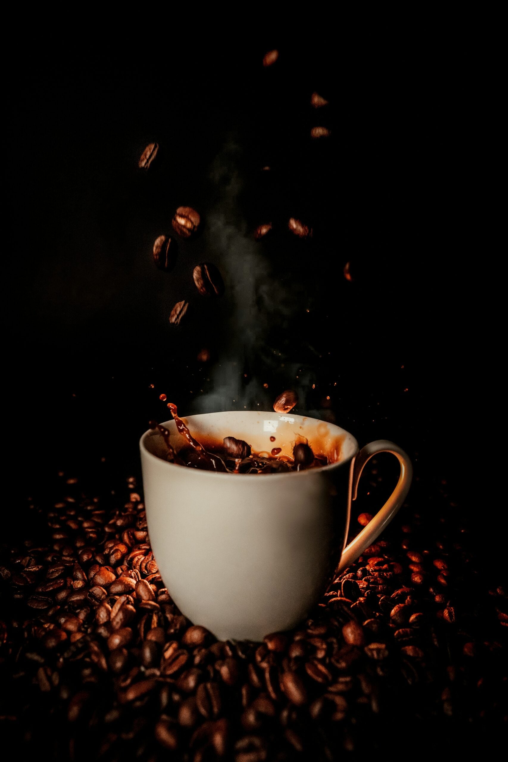 A white coffee cup sits on a pile of coffee beans, with coffee splashing inside and beans falling into the cup against a dark background. Steam rises from the hot coffee.