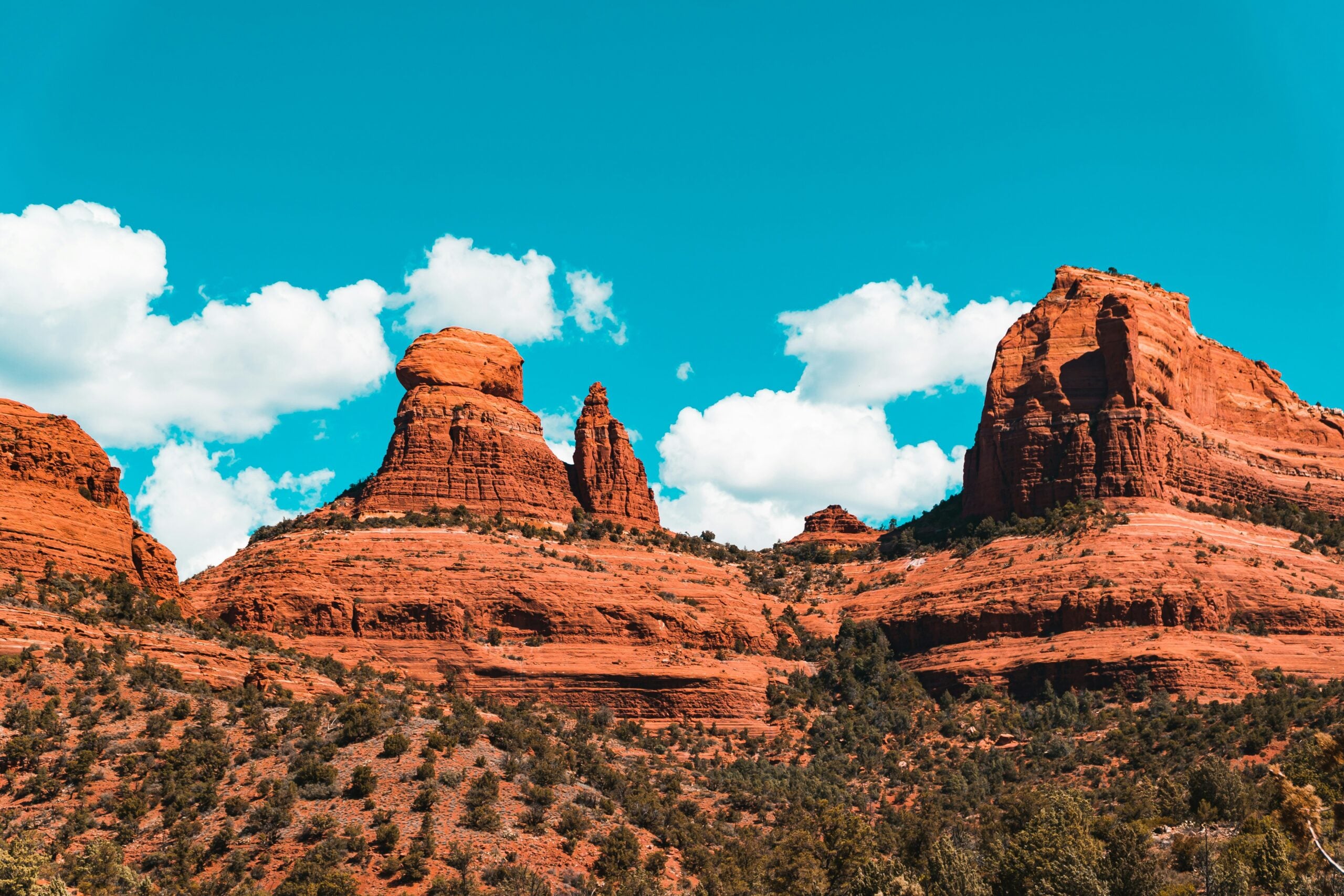 Red rock formations rise against a bright blue sky with scattered white clouds, surrounded by green vegetation at their base.