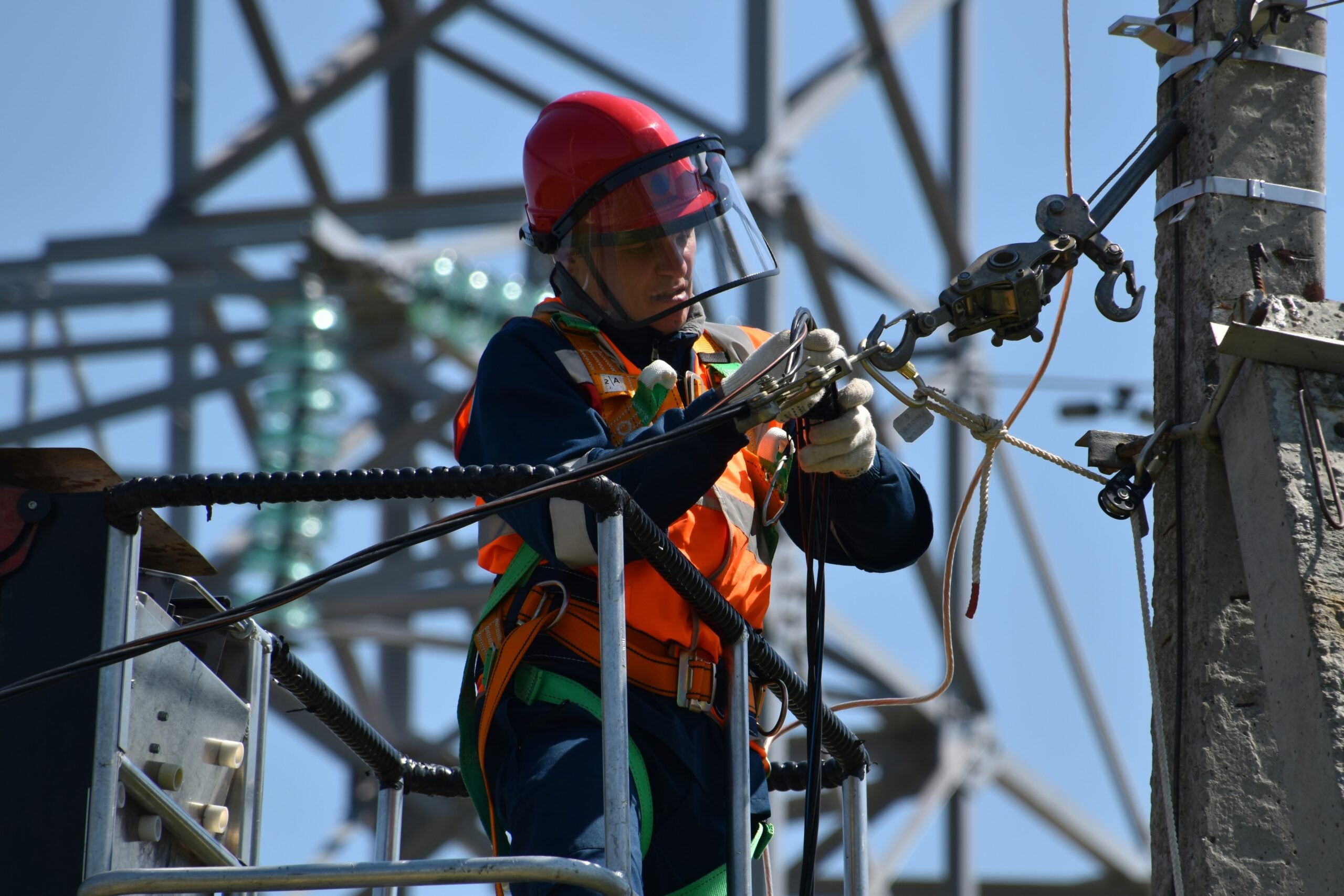 A utility worker wearing safety gear and a red helmet is working on electrical cables atop a lift near a utility pole, with a power line structure visible in the background under a clear sky.