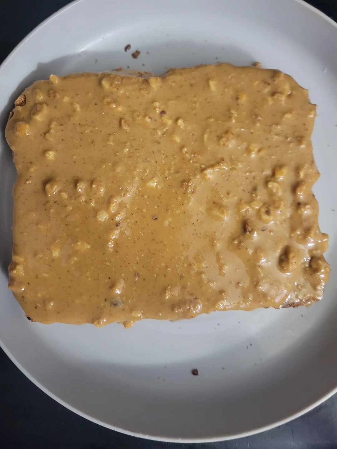 A slice of bread covered with chunky peanut butter sits on a white plate.