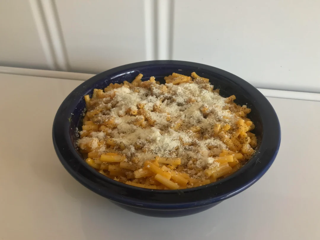 A blue bowl filled with macaroni and cheese, topped with grated Parmesan cheese and a sprinkle of black pepper, sits on a white surface against a white background.