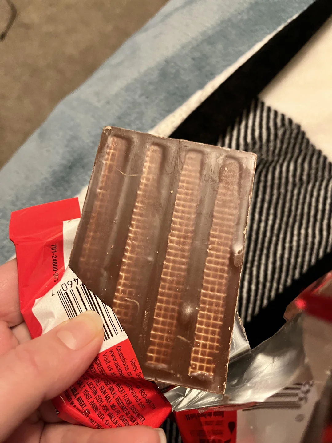 A hand holds a chocolate bar partially unwrapped, revealing that the chocolate is solid with visible outlines of where Kit Kat fingers would normally be separated, but without distinct breaks.