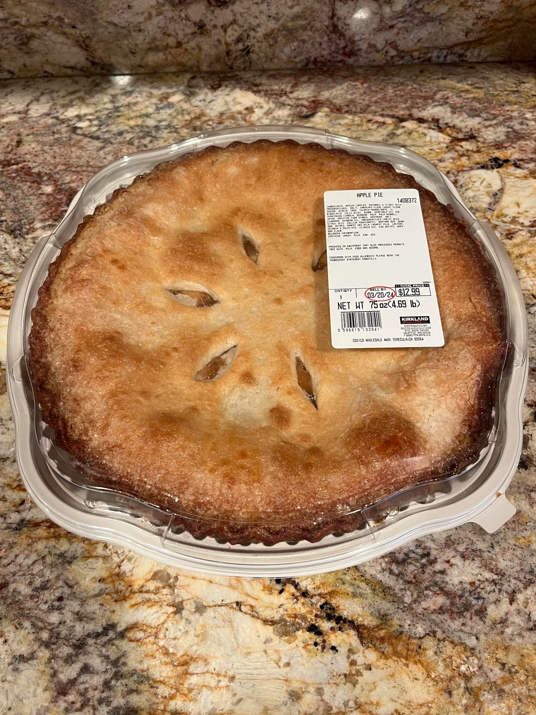 A whole apple pie in a clear plastic container sits on a marbled countertop. A white label with product and pricing information is attached to the container’s lid. The pie has a golden-brown crust with several slits on top.