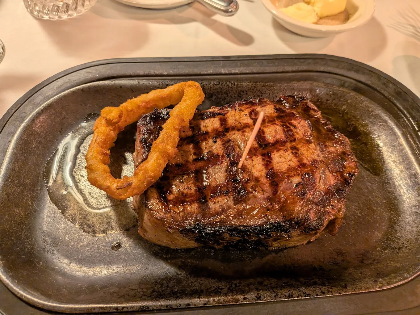 A grilled steak with visible char marks is served on a metal plate, topped with a single onion ring. A dish of butter and utensils are visible in the background on a white tablecloth.