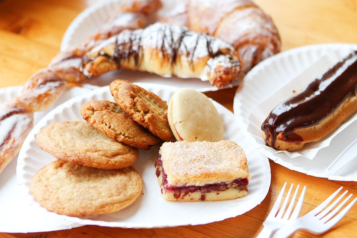 Assorted pastries and cookies, including a chocolate éclair, macaroon, cinnamon twist, and fruit bar, arranged on white paper plates on a wooden table with plastic forks nearby.