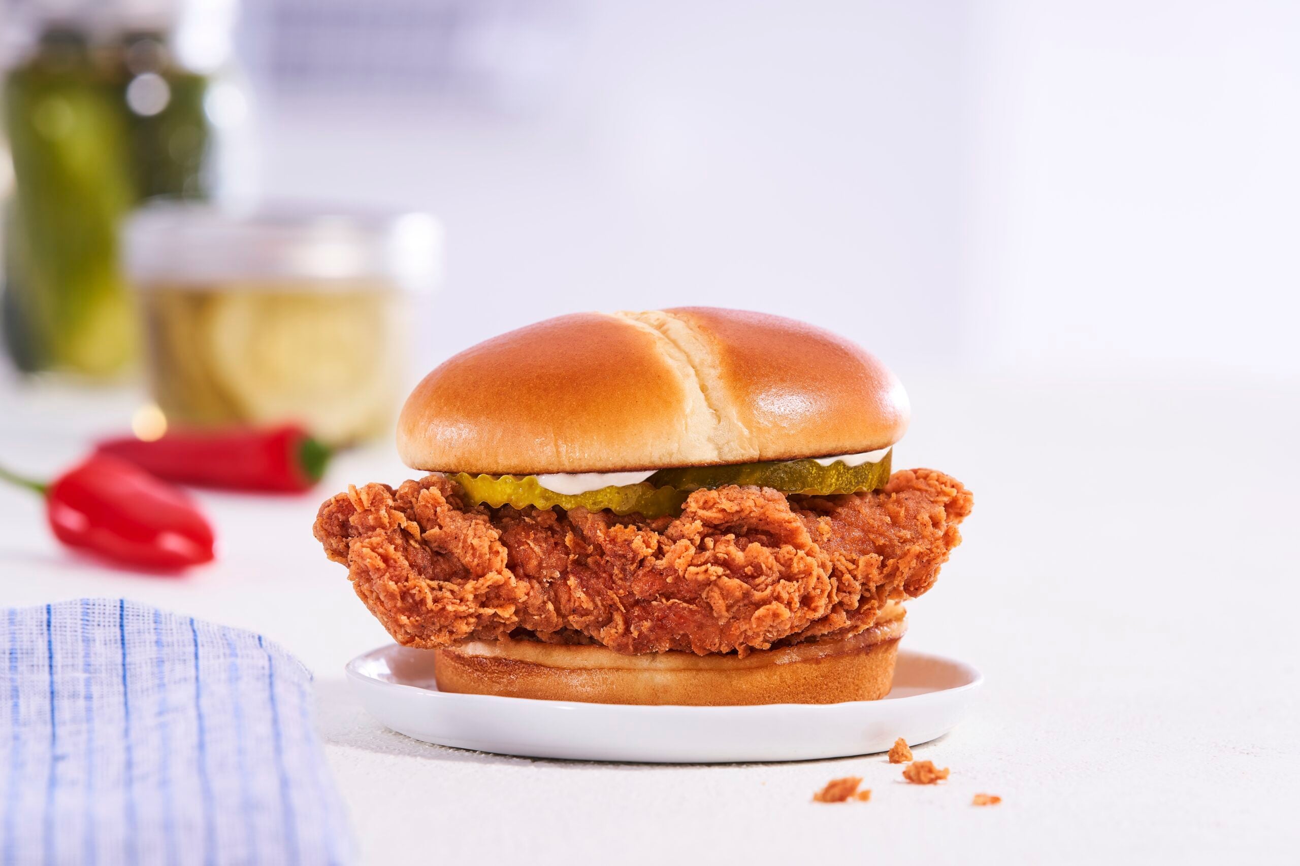 A crispy Chicken Sandwich with pickles and mayonnaise on a soft, toasted bun, served on a white plate with a blurred background and a red chili pepper nearby.