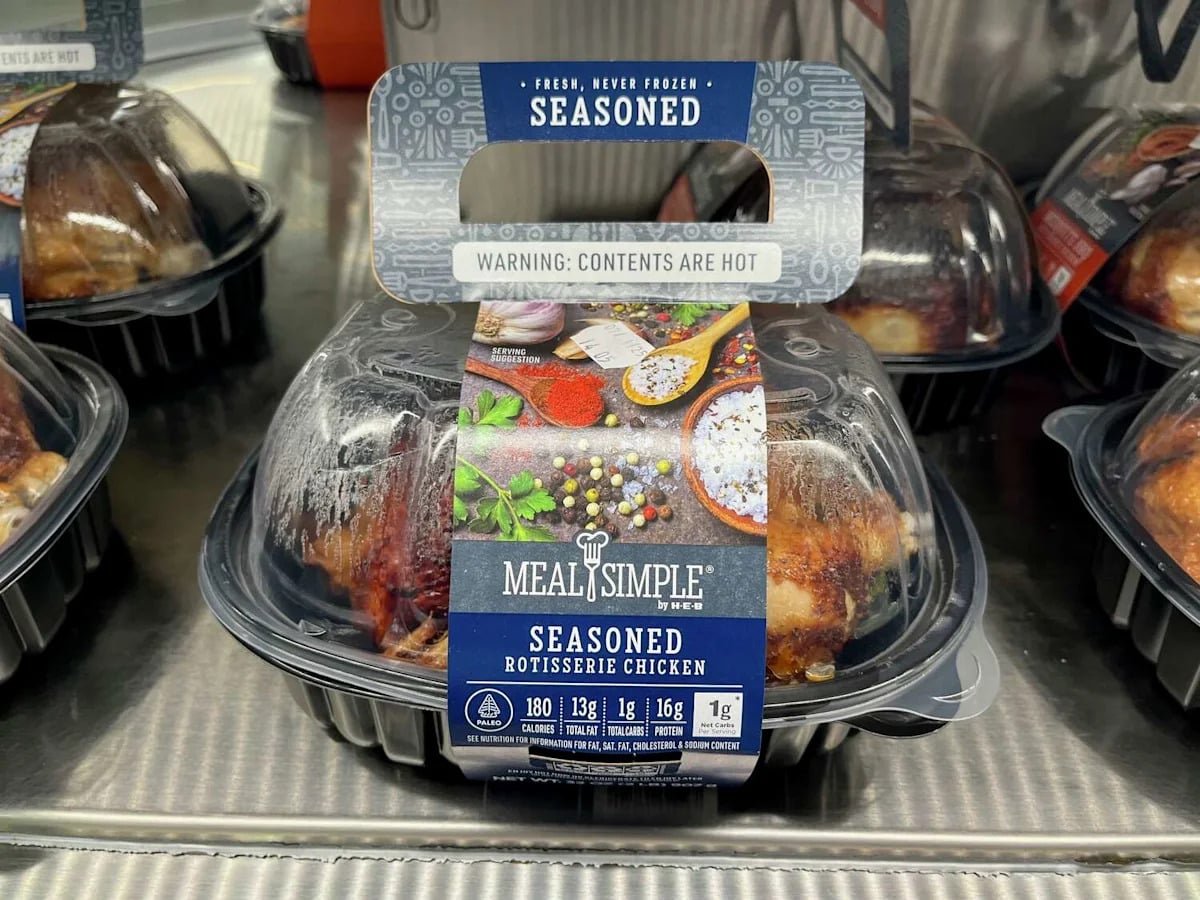 A packaged Rotisserie Chicken labeled "Meal Simple Seasoned Rotisserie Chicken" sits on a metal counter, warning that contents are hot, with images of spices and herbs highlighting the savory flavors.