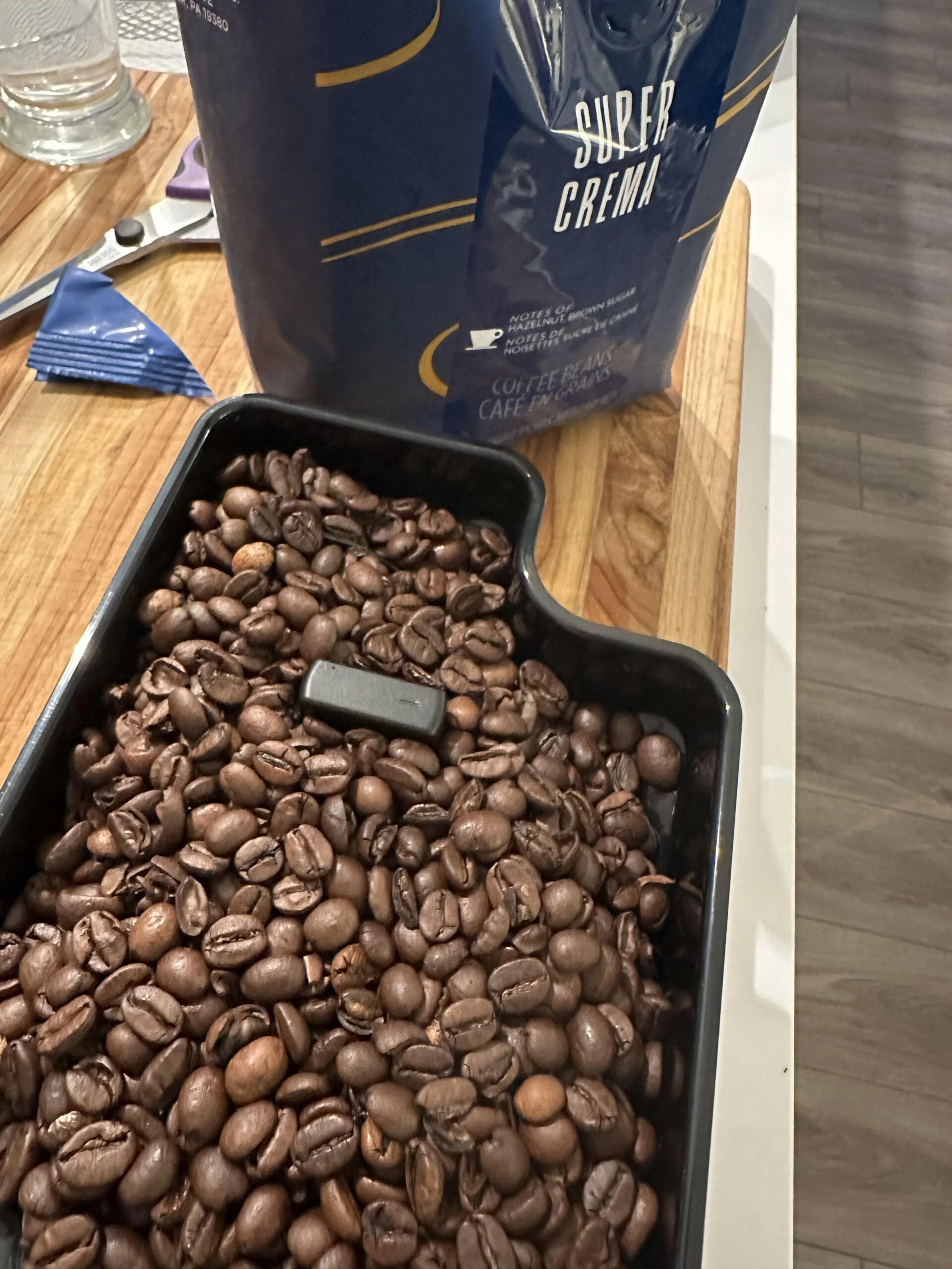 A tray filled with dark roasted coffee beans sits on a wooden kitchen counter next to an open bag labeled "Super Crema," one of the popular coffee brands. The partially visible bag adds a touch of authenticity to the cozy scene.