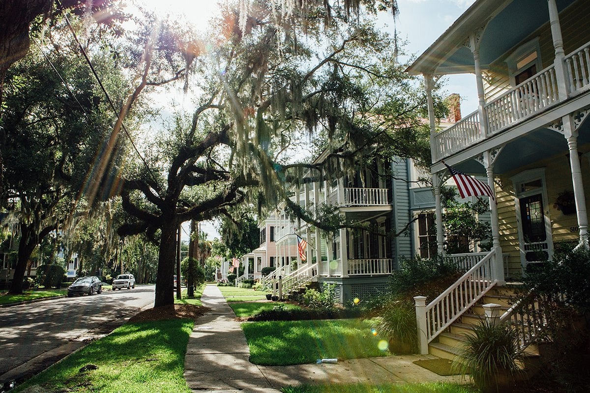 Sunlight filters through large, moss-draped oak trees onto a quiet street lined with charming, colorful houses—hallmarks of Unique American Towns—each porch decked with American flags, creating a warm, inviting neighborhood atmosphere.