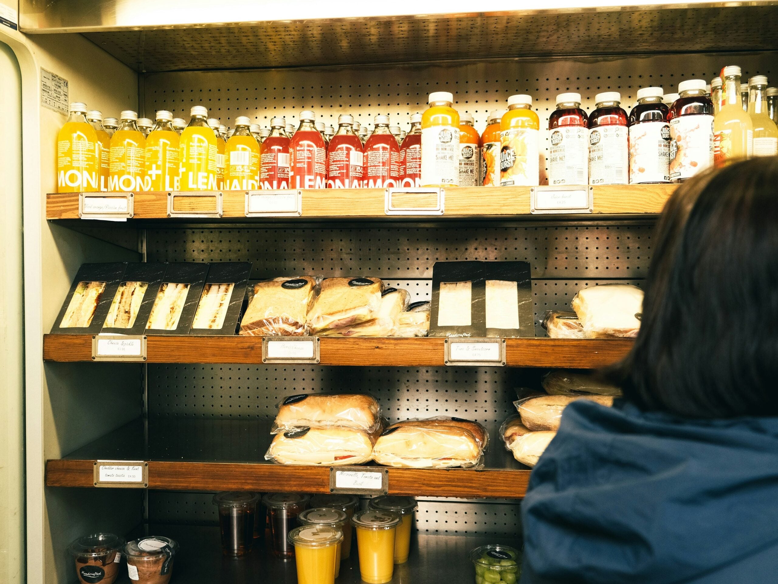 A refrigerated display case with bottled drinks on the top shelves and neatly packaged sandwiches, salads, and snacks on the lower shelves. A person stands in the foreground, partially visible from behind.