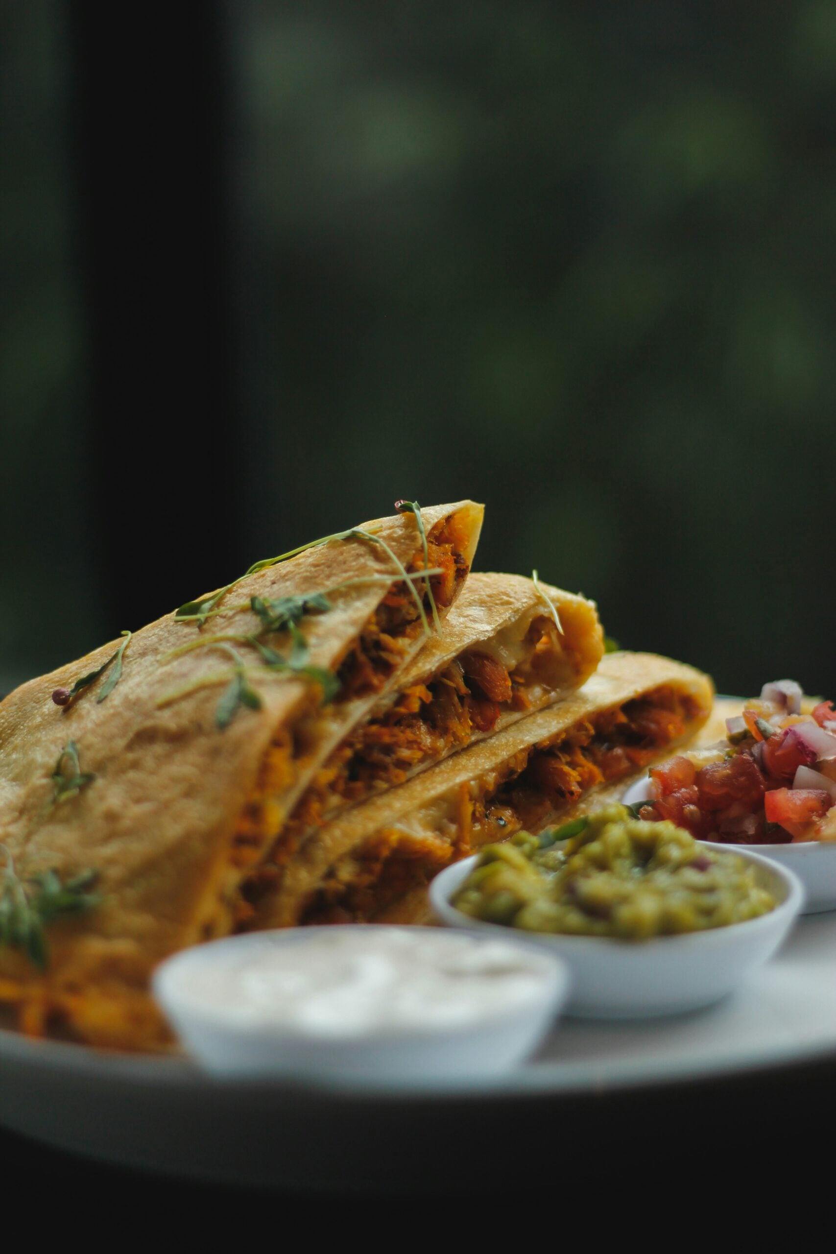 A close-up of a quesadilla cut into wedges, garnished with herbs and served on a white plate with small bowls of sour cream, guacamole, and salsa. The background is blurred and green.