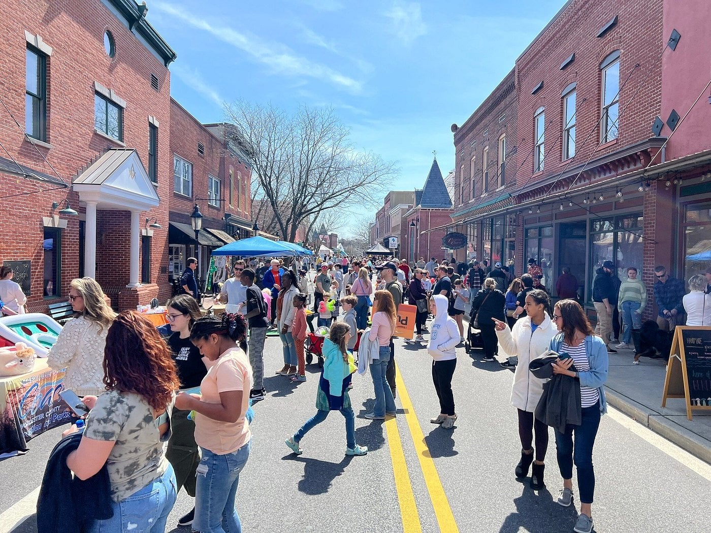 A bustling street fair in one of those Unique American Towns, where crowds walk, browse vendor booths, and socialize between red brick buildings under a clear blue sky on a sunny day.