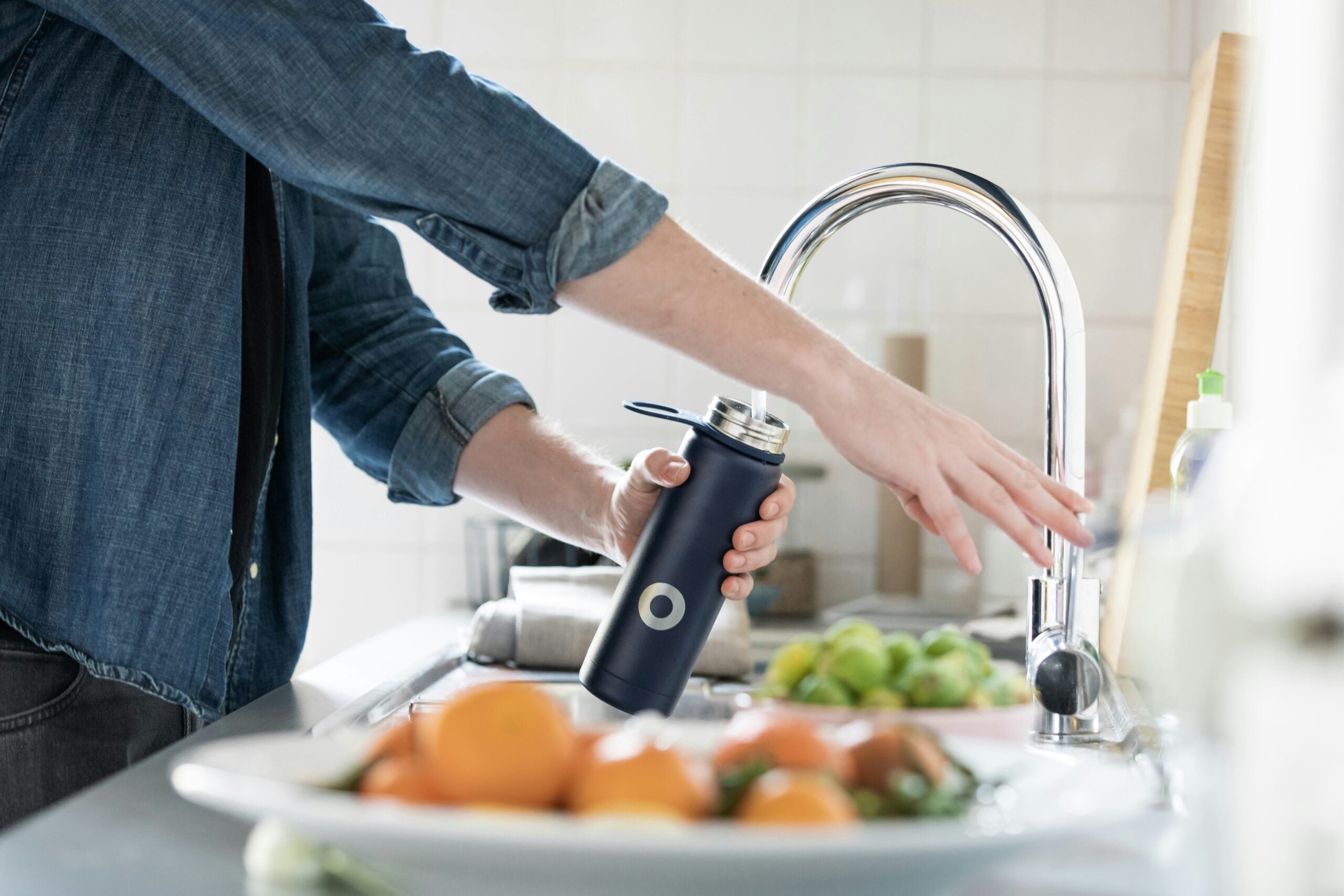 A person in a denim shirt fills a dark blue water bottle at a kitchen sink, with fresh fruits and vegetables visible in the foreground on a white plate.