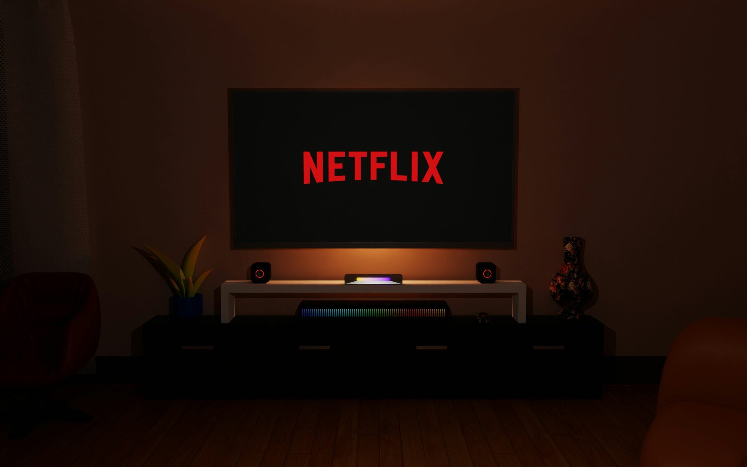 A dimly lit living room with a large TV displaying the red Netflix logo, speakers on each side, a plant, and a decorative vase on a white TV stand. Warm lighting creates a cozy atmosphere.