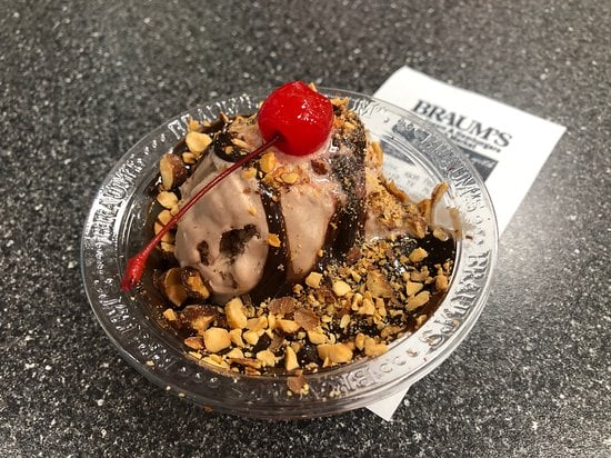 A chocolate sundae topped with chopped peanuts, hot fudge, and a maraschino cherry, served in a clear plastic bowl on a dark countertop with a Braum’s napkin underneath.
