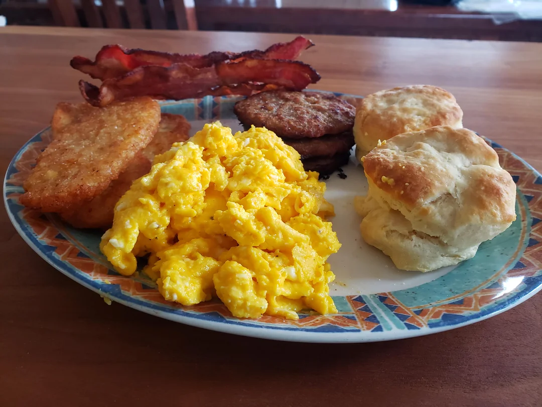 A plate of scrambled eggs, two hash browns, two sausage patties, two strips of bacon, and two biscuits on a patterned dish, set on a wooden table.
