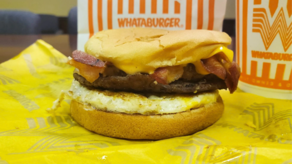 A Whataburger fast-food breakfast sandwich with bacon, egg, beef patty, and cheese in a bun sits on yellow wrapping paper, with Whataburger-branded cups and packaging in the background.