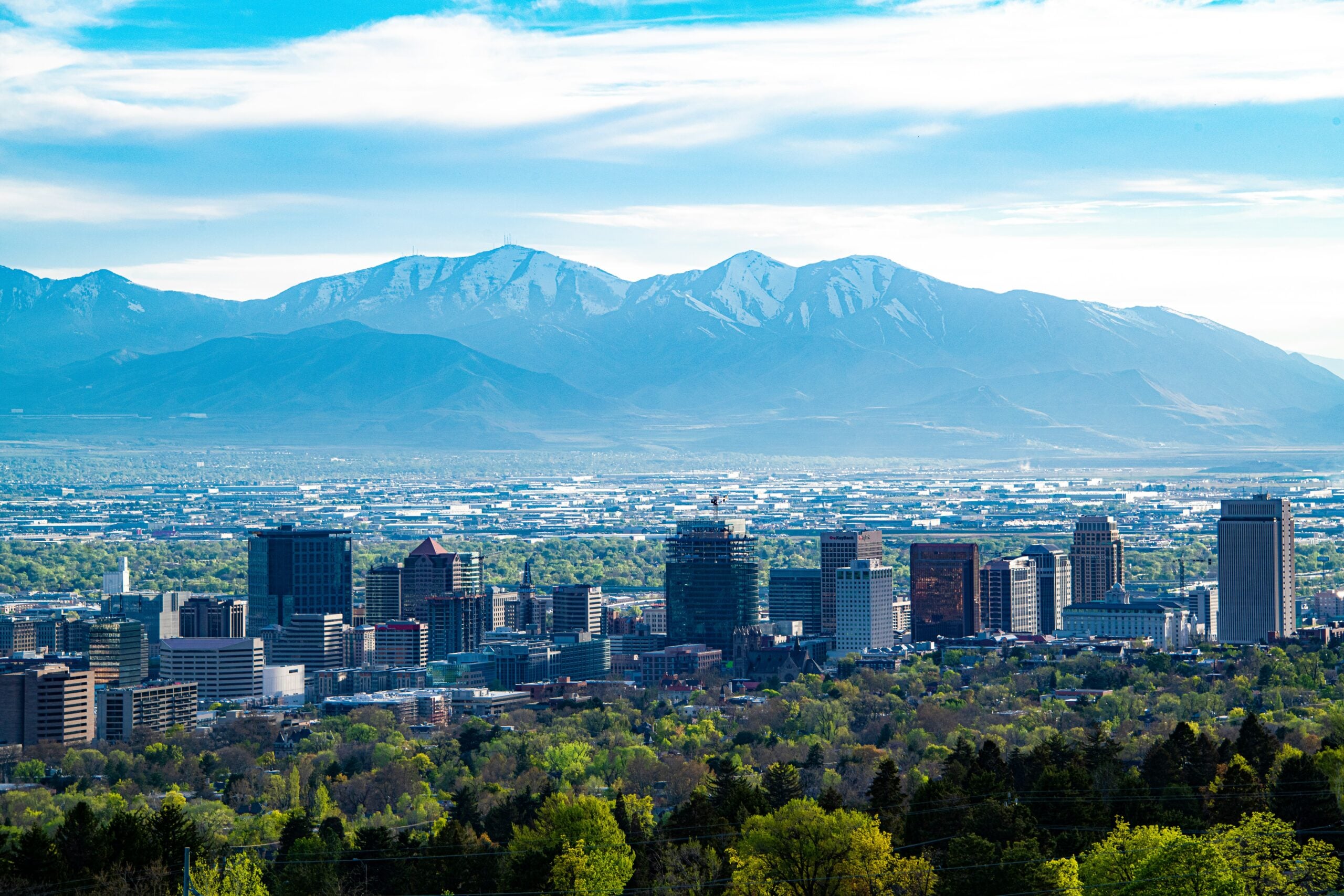 A city skyline with tall buildings in the foreground, lush green trees below, and snow-capped mountains rising in the background under a blue sky with light clouds.