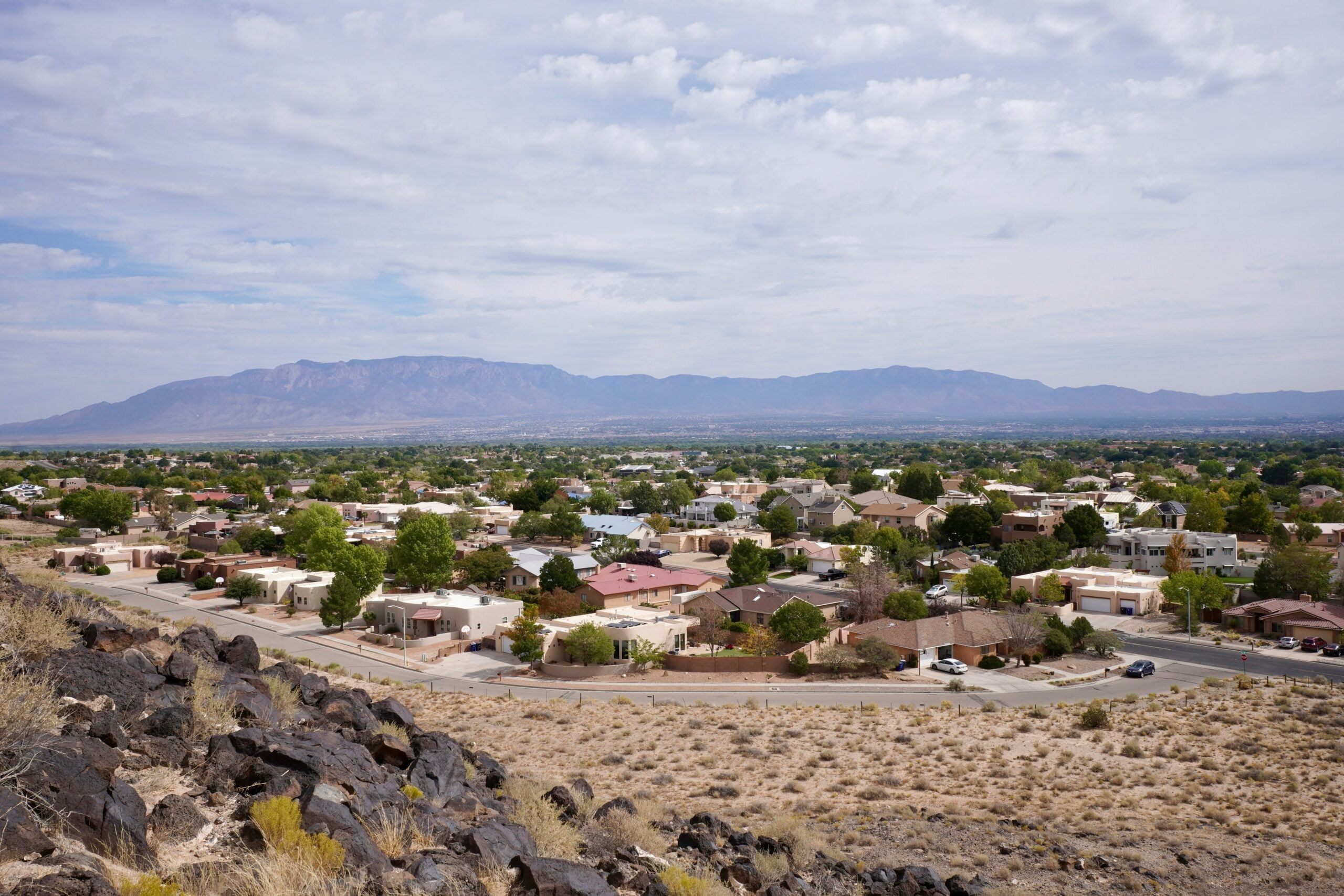 A suburban neighborhood with single-story houses, green trees, and streets, set against distant mountains under a partly cloudy sky. Rocky terrain and dry grass appear in the foreground, reflecting the need for Cost of Living Relief in this peaceful setting.