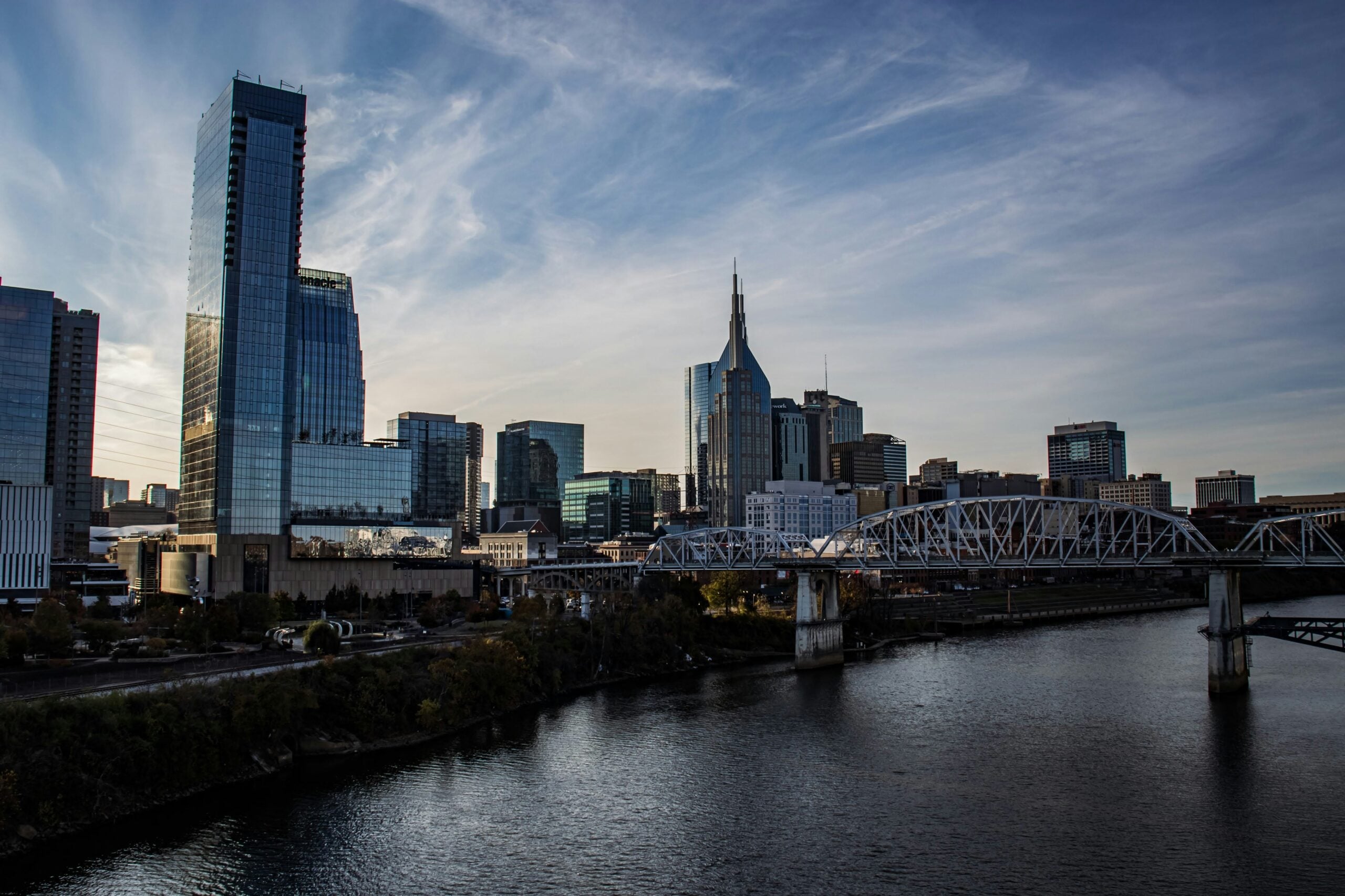 A city skyline with modern high-rise buildings, a distinctive tower with a spire, and a large bridge crossing a river under a partly cloudy sky at sunset.