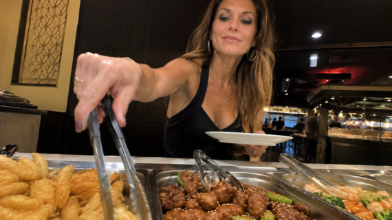 A woman holding a white plate uses tongs to serve herself food from an All-You-Can-Eat Buffet, with fried items and saucy dishes visible in metal trays. She is smiling and wearing a sleeveless black top.