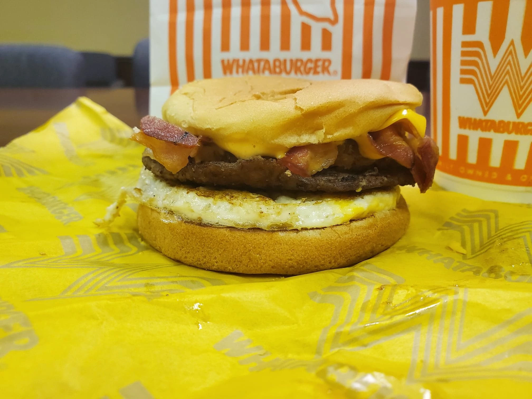 A Whataburger breakfast sandwich with bacon, egg, cheese, and a beef patty on a bun, sitting on yellow Whataburger-branded paper with a Whataburger cup and bag in the background.