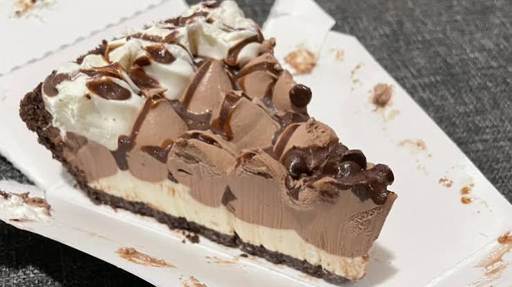 A slice of layered chocolate and vanilla cream pie with a chocolate crust, topped with whipped cream and chocolate drizzle, served on a white paper tray.