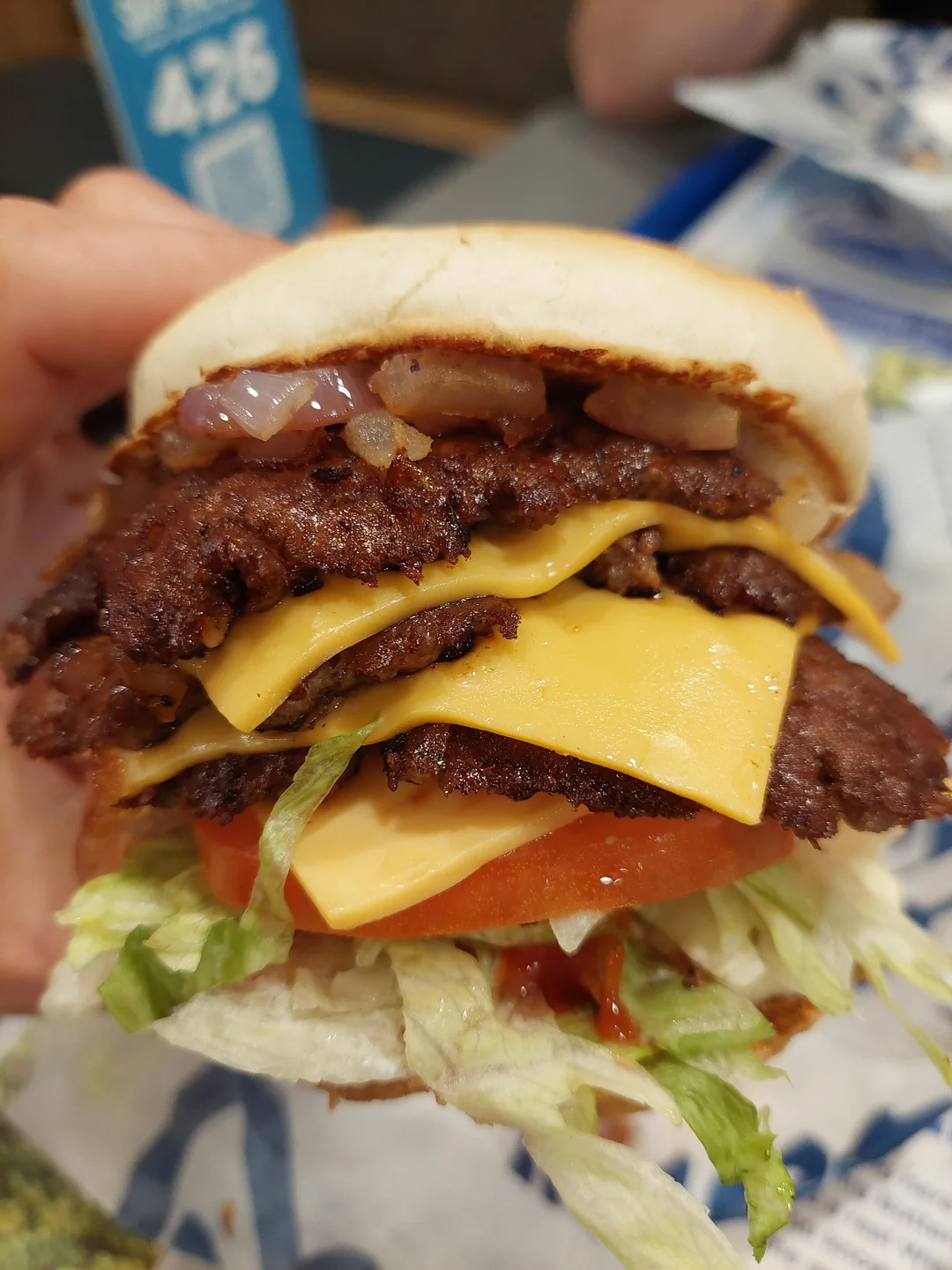 A close-up of a hand holding a cheeseburger with two beef patties, two slices of cheddar cheese, chopped onions, lettuce, tomato, and ketchup on a hamburger bun.