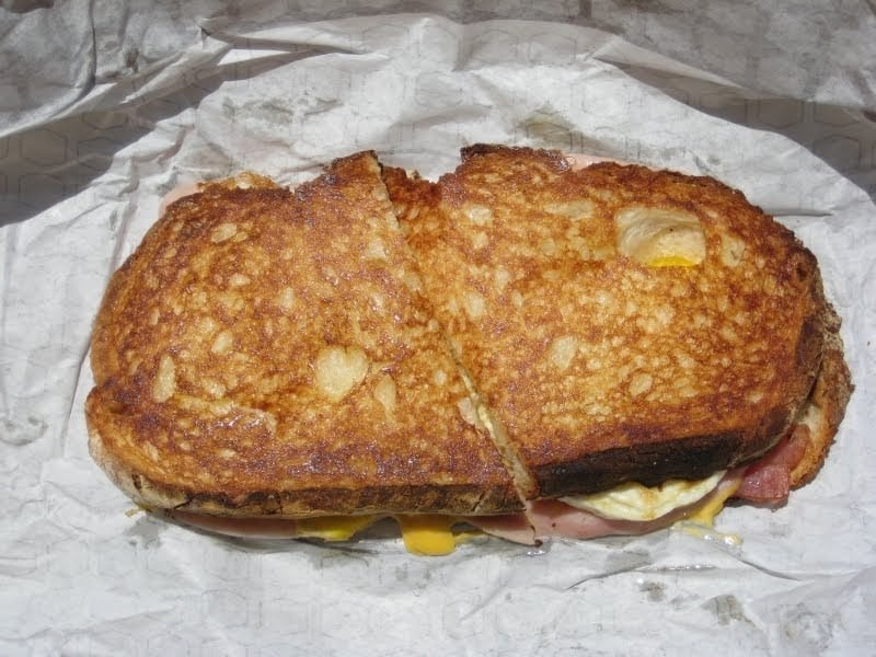 A toasted sandwich with a golden-brown crust, cut in half and placed on a sheet of crumpled white paper. Melted cheese and ham are visible between the bread slices.