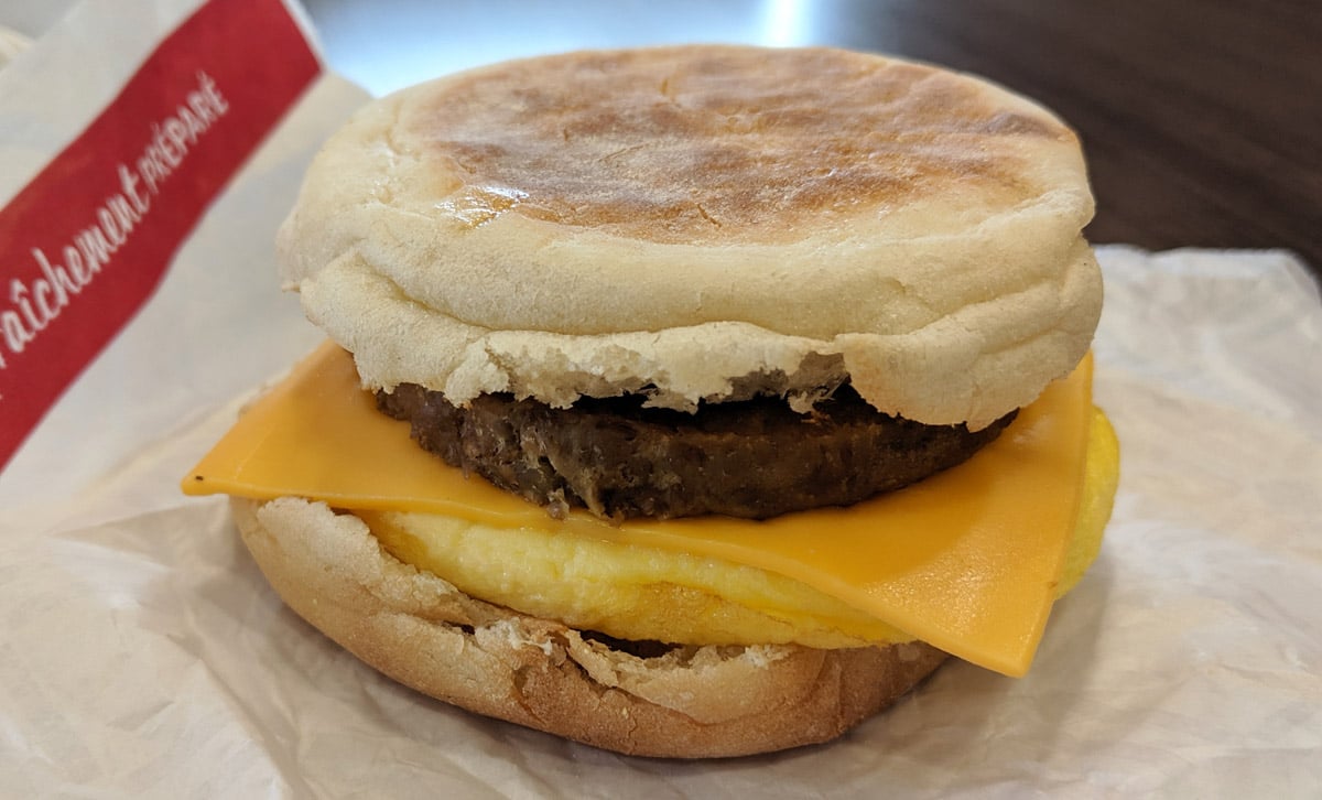 A breakfast sandwich with an English muffin, a slice of cheddar cheese, a sausage patty, and a folded egg, placed on a white wrapper with part of a red label visible in the background.