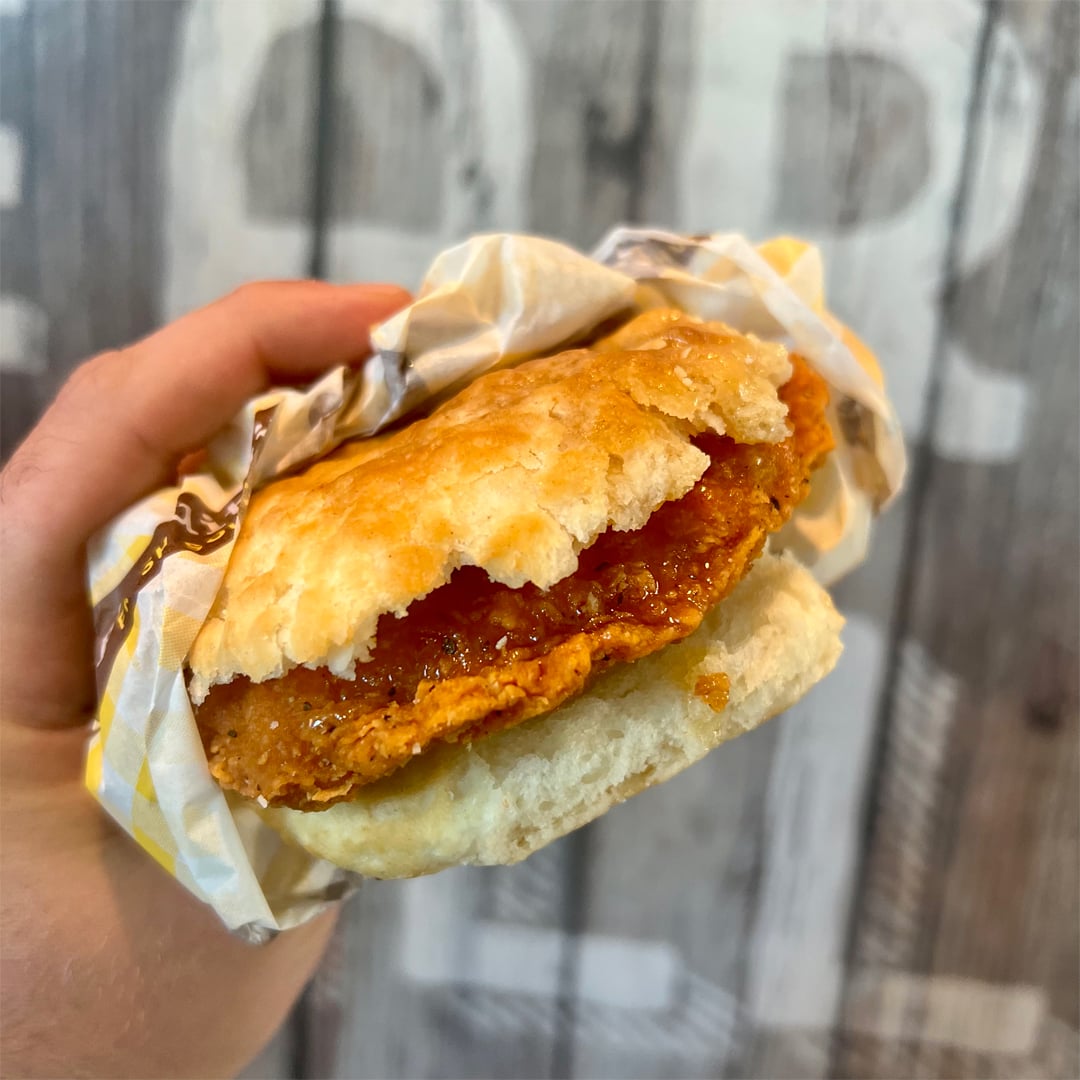 A hand holds a biscuit sandwich filled with a crispy fried chicken fillet, partially wrapped in yellow and white paper, against a blurred wooden background with large, faded letters.