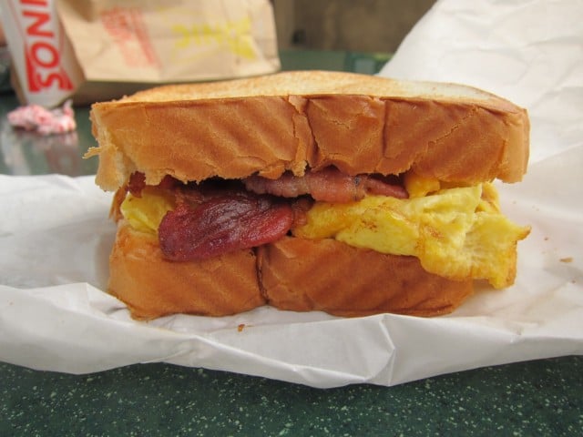 A breakfast sandwich with scrambled eggs and crispy bacon between two slices of toasted bread, placed on white paper.