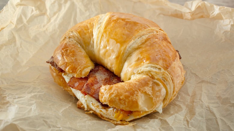 A flaky croissant sandwich filled with crispy bacon and a fried egg, resting on crumpled brown parchment paper.