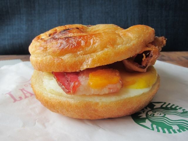 A close-up of a breakfast sandwich with bacon, egg, and cheese inside a golden, flaky croissant bun, placed on a Starbucks-branded paper wrapper.