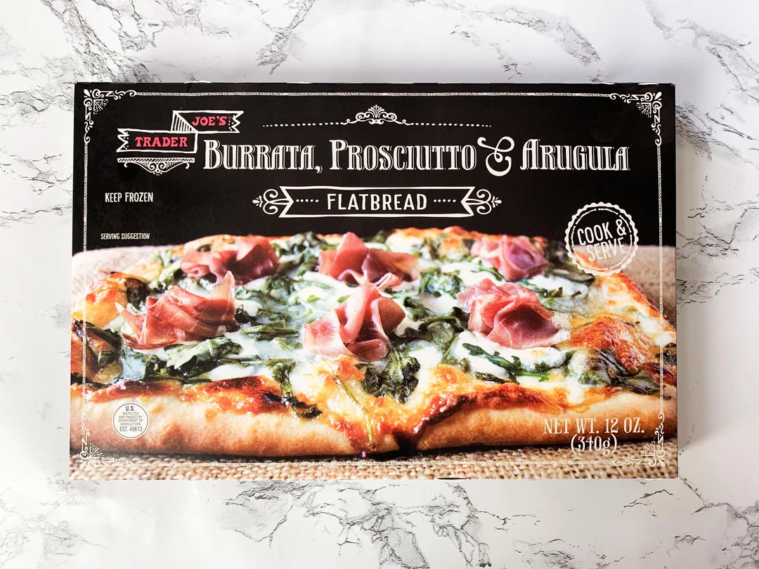 A box of Trader Joe’s Burrata, Prosciutto & Arugula Flatbread, featuring an image of the flatbread topped with cheese, prosciutto, and greens on a marble surface. The box states “Cook & Serve” and 12 oz.