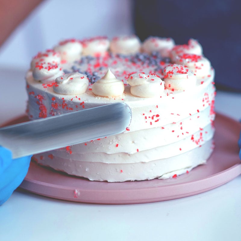 A person wearing blue gloves is slicing a white frosted cake decorated with red sprinkles and piped cream on top, placed on a pink plate.