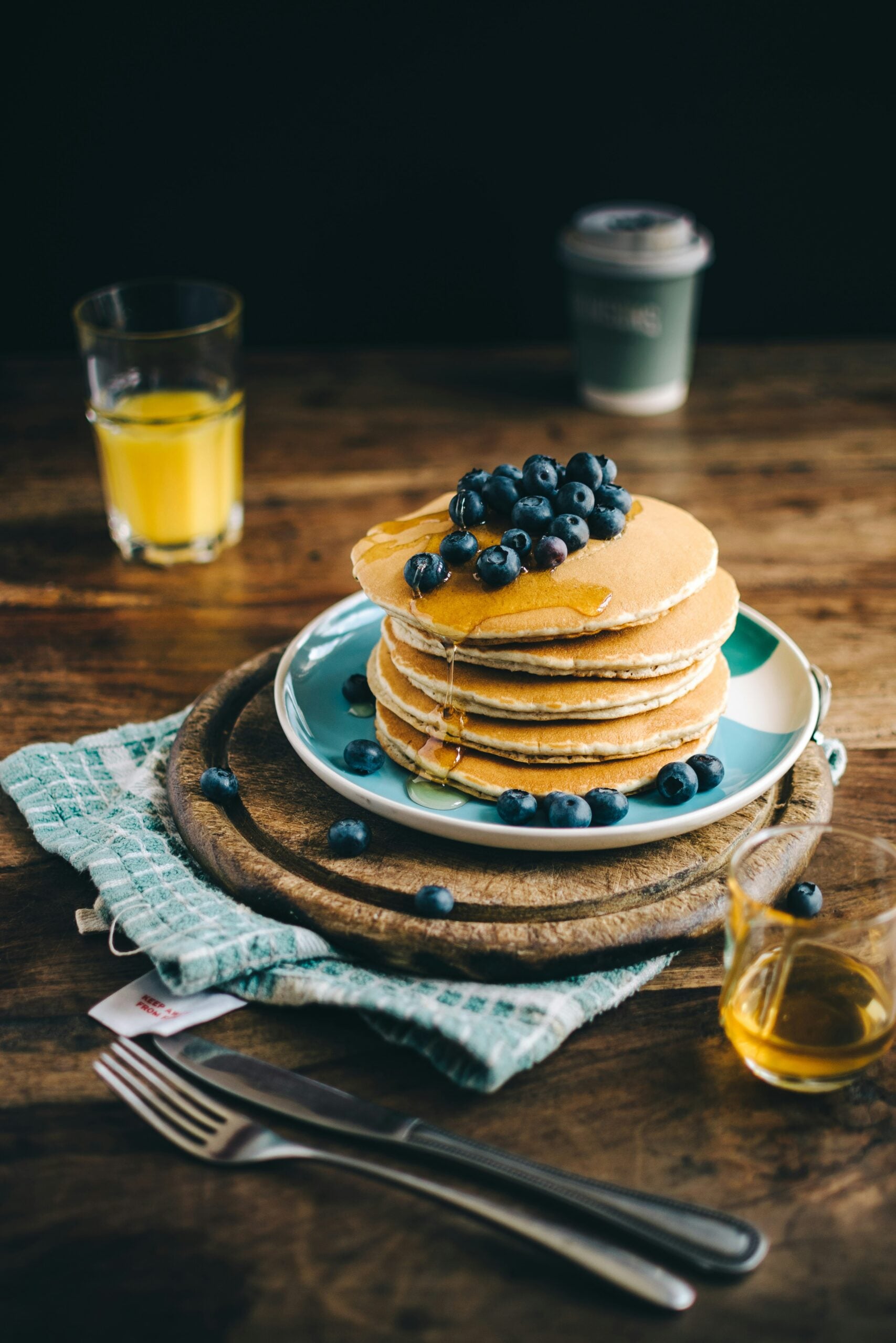 A stack of pancakes topped with blueberries and syrup sits on a plate, with a fork and knife nearby. A glass of orange juice, a coffee cup, and a small pitcher of syrup are on a rustic wooden table.