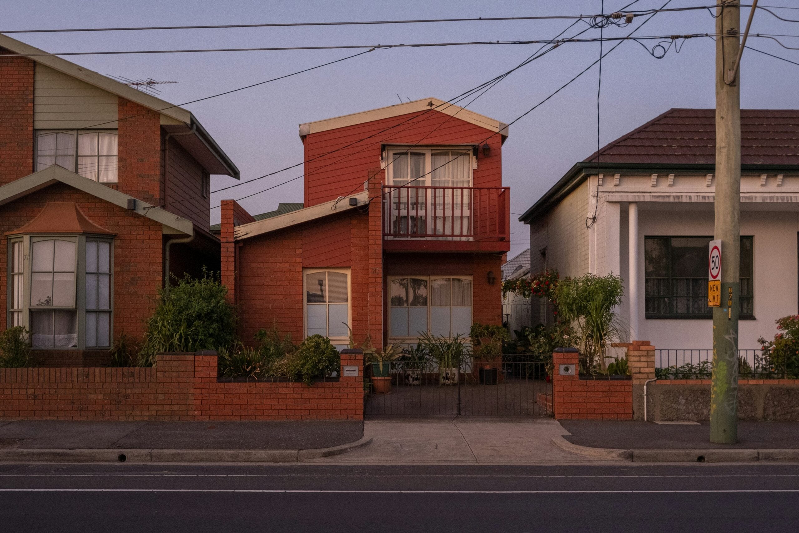 A two-story red house with a balcony is situated between a brick house and a white house. Potted plants decorate the front yard, and power lines run overhead. A sidewalk and road are in the foreground.