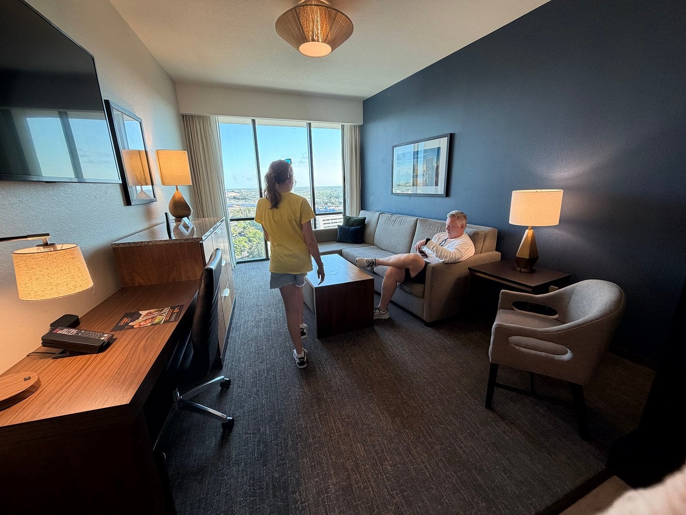 A woman walks into a modern hotel room with dark walls and large windows, while a man sits on a gray sofa using a phone. Showcasing city views and sleek decor, this space reflects the elevated standards of top Hotel Brands 2026.