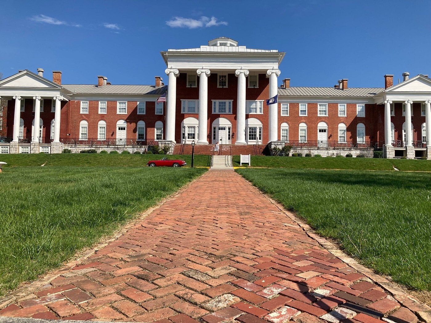 A red brick pathway leads to a large, red brick building with white columns and trim, set against a bright blue sky—an inviting sight reminiscent of Small-Town Luxury Hotels. A small red car is parked on the driveway in front of the building.