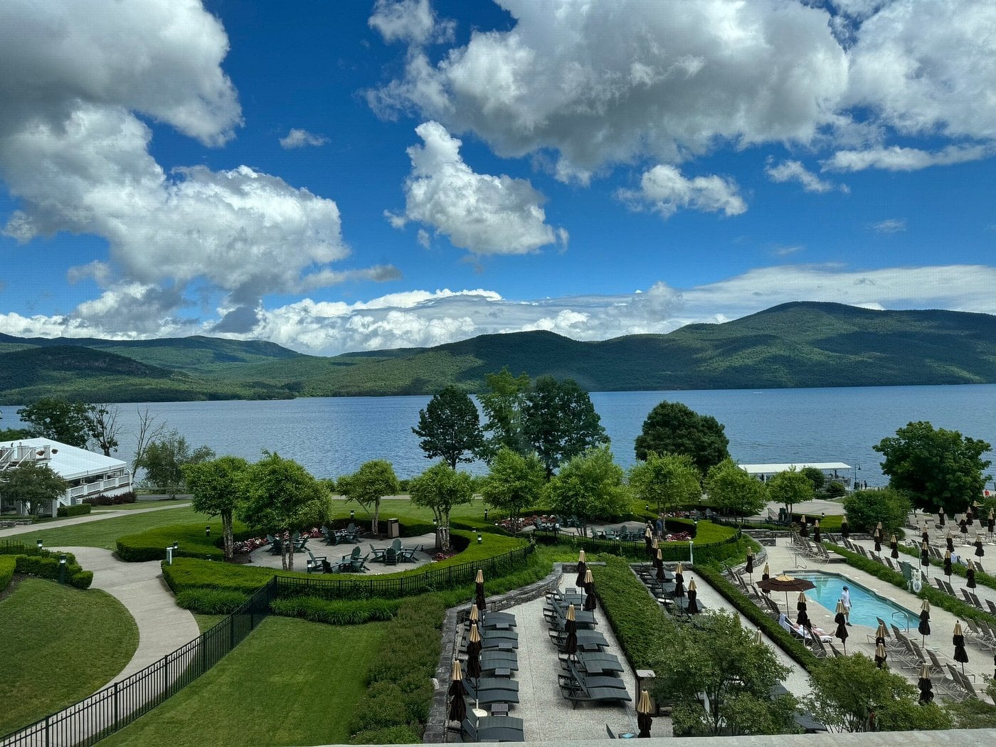 A scenic view of a lake surrounded by green hills under a partly cloudy sky, with a pool, lounge chairs, and landscaped gardens in the foreground. Guests at this Small-Town Luxury Hotel are relaxing by the pool and strolling through the grounds.
