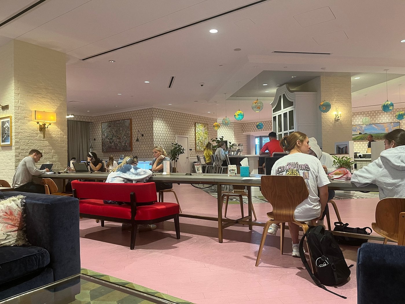 People sit and work on laptops at a long communal table in a modern, brightly lit lounge with pink floors—found in some Small-Town Luxury Hotels—surrounded by colorful decor and wall art as others chat or relax.