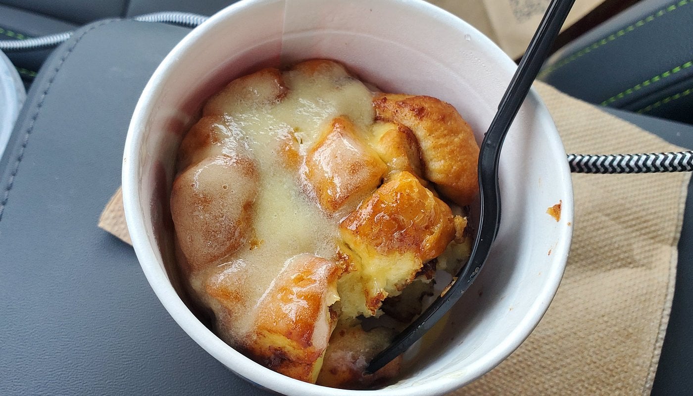 A white paper cup filled with cinnamon roll bites covered in icing, with a black plastic fork sticking into the dessert. The cup is placed on a napkin inside a car.