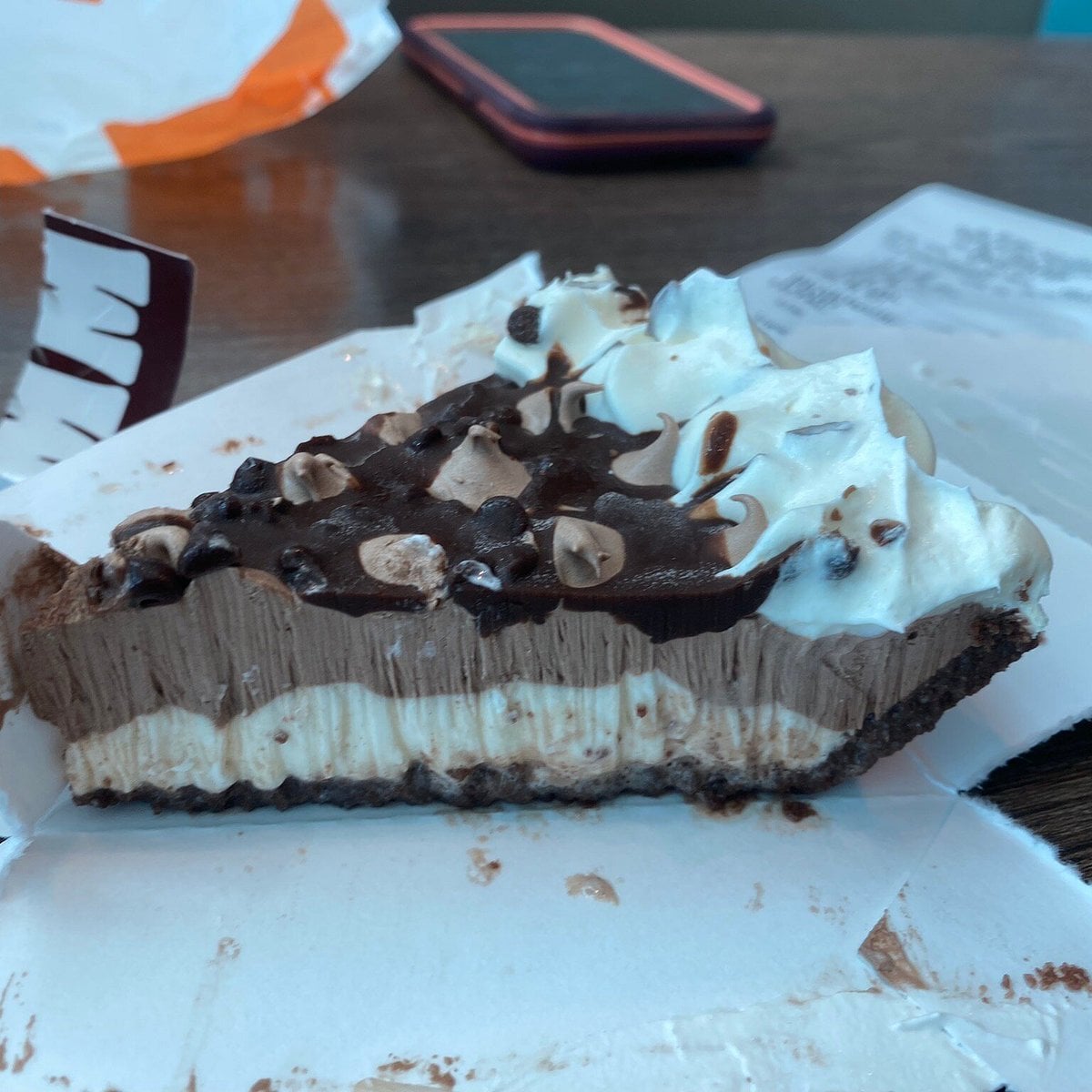 A slice of chocolate and cream pie with a chocolate crust, topped with whipped cream, chocolate chips, and chocolate drizzle, resting on white paper with a blurred cellphone and other items in the background.