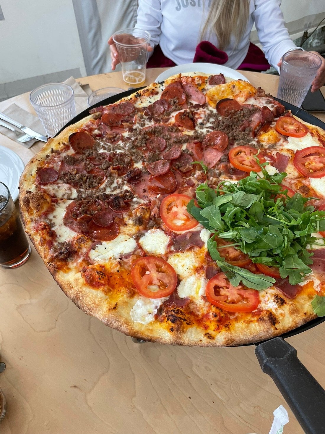 A large pizza with four distinct sections—sliced tomatoes and arugula, pepperoni, tomato slices, and ground meat—on a wooden table with drinks and cutlery. Perfect for sharing at road trip food stops, with two people partially visible in the background.