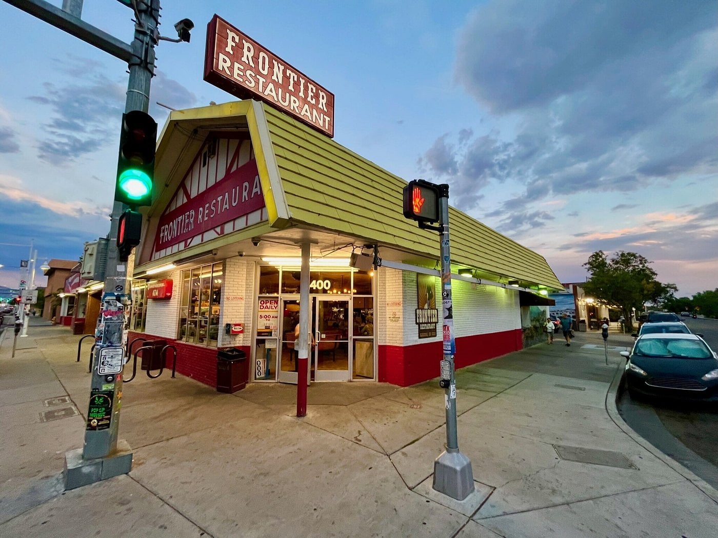 A yellow and red restaurant called Frontier Restaurant sits on a street corner at sunset—a classic road trip food stop—its sign glowing as cars line the curb and people gather near the entrance under a green traffic light.
